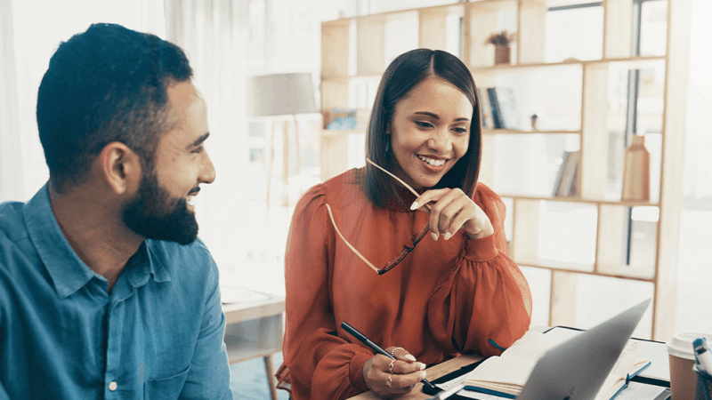 Woman sits next to coworker, looking at computer screen but engaged in conversation