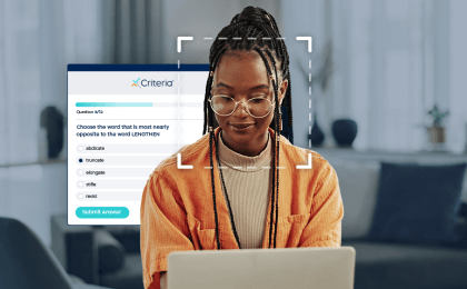 Young woman sits in front of a laptop taking an assessment - overlayed, there is a facial recognition square and Criteria assessment UI in the background, indicating that she is taking her assessment with proctoring capabilities.